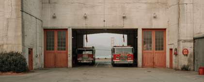 Two fire trucks are parked inside an open garage of a fire station, with the Golden Gate Bridge visible in the background through the open doors.