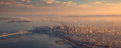Aerial view of San Francisco at sunset, showing the skyline, Bay Bridge, waterfront piers, and distant hills under a partly cloudy sky.