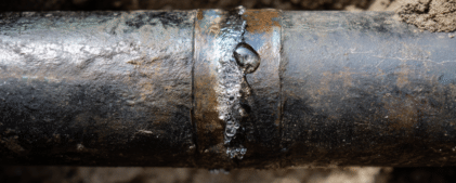A close-up of a metal pipe with water leaking and bubbles forming at a joint, surrounded by dirt.