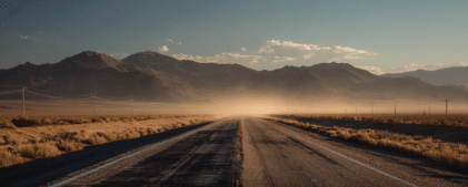 A straight, empty road stretches into the distance with dust rising ahead, flanked by dry terrain and mountains under a clear sky.