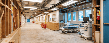 A hospital hallway under renovation with exposed walls, construction materials, hospital beds, and medical equipment along the corridor.
