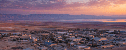 A small desert town with scattered buildings and palm trees sits near a reflective lake and distant mountains under a colorful sunset sky, recently experiencing the effects of the Brawley earthquake swarm 2026.