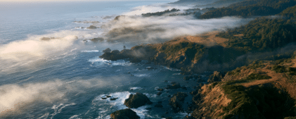 Aerial view of a rocky coastline with waves crashing, coastal cliffs, fog rolling in from the ocean, and forested hills—an awe-inspiring landscape shaped by tectonic forces like the Cascadia San Andreas earthquake.