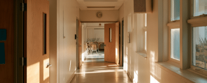 Empty school hallway with sunlight streaming through windows, open wooden doors, a wall clock, and a classroom at the end with chairs stacked on desks—an atmosphere of calm enhanced by the reassurance of a school earthquake early warning system.