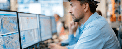A man wearing a white hard hat and blue shirt works at multiple computer monitors displaying technical data in an office setting for earthquake early warning system facilities.